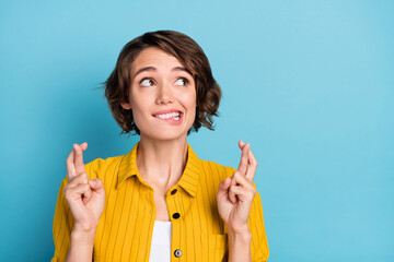 Photo of nervous hopeful lady bite lip crossed fingers look empty space wear yellow shirt isolated blue color background