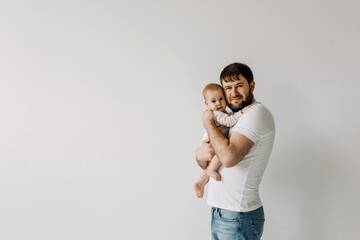 Father holding baby in arms, smiling, on white wall background, dressed in white clothes.