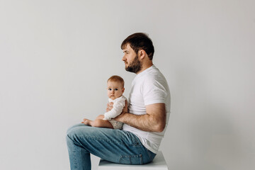 Father sitting, holding baby on his laps, on white wall background.
