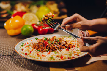 Rice food in plate with fresh vegetable ingredients behind and woman's hands holding fork with meat