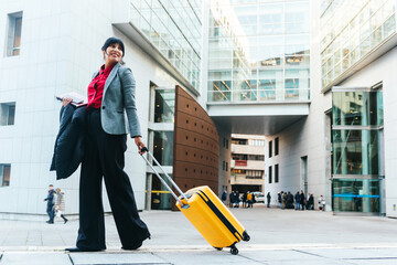 Latin business woman with yellow suitcase walks through the financial center of a city during a business trip.