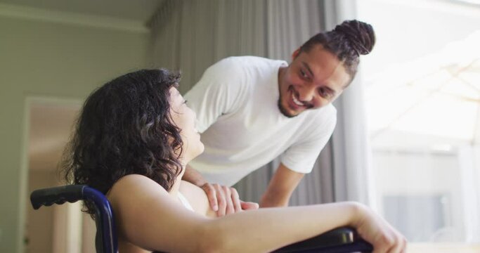 Happy biracial woman in wheelchair and smiling male partner with dreadlocks talking at home