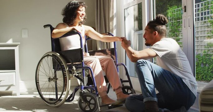 Happy biracial woman in wheelchair with smiling male partner kissing hand in sunny living room
