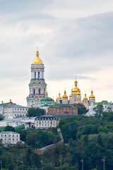 The bell tower of the Pechersk Lavra and the golden domes of the monastery on the slopes of the Kyiv hills on a cloudy summer day.