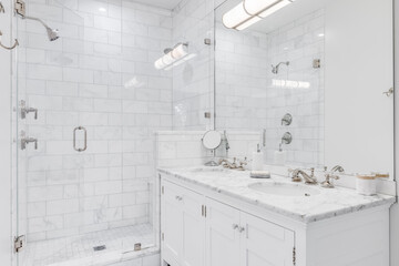 A luxurious white bathroom with a double vanity and bronze faucets, a white subway tile shower, and...