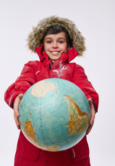 Cheerful European boy in bright red snowy down jacket with hood, holding huge globe in his hands, smiles looking at camera, isolated on white background. Tourism, geography, planet exploration concept