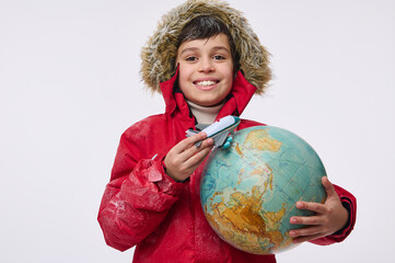 Fototapeta premium Around the world travel and winter tourism concept. Adorable elementary aged boy in warm bright red down jacket with hood holding an airplane toy model and globe, imitating the flight around the Earth