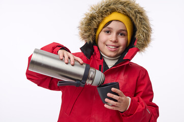 Horizontal portrait of a cute school-age boy winter traveler adventurer pouring hot drink from thermos into mug smiling toothy smile looking at camera isolated on white background with copy space - obrazy, fototapety, plakaty