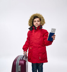 Isolated portrait on a white background of a handsome preteen Caucasian boy in bright red parka with eco further hood, looking at camera posing with a suitcase and holding a passport and boarding pass © Taras Grebinets