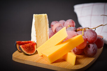 Side view of cheeseboard with served on dark background. Hard and soft French cheese pieces with grape and fig cuts. White mould cheese on plate. Appetizers on wooden board. Fruits and berries
