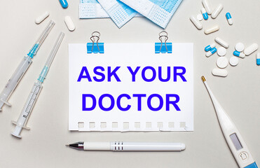 On a light gray background, blue medical masks, syringes, an electronic thermometer, pills, a pen and a notebook with the inscription ASK YOUR DOCTOR . Medical concept