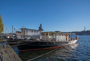 Obraz premium Various boats at the pier Norrmälarstrand at the bay Riddarfjärden in the district Kungsholmen, the tower of the Town City Hall a sunny color full autumn day in Stockholm