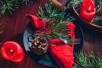 Festive Christmas dinner table setting decorated with fir branches, red candles, cones, ornaments.