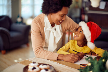 Happy African American mother talks to her daughter who is writing letter to Santa for Christmas.