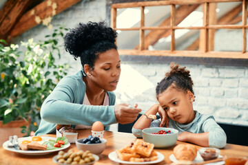 African American mother feeds her small daughter who is refusing to eat her breakfast at dining...