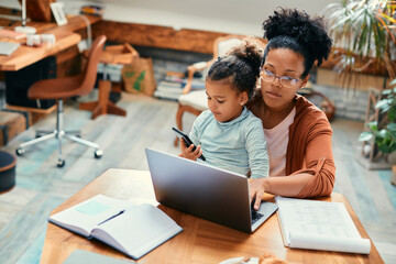 African American stay at home mother works on laptop while daughter is sitting on her lap and using smart phone.