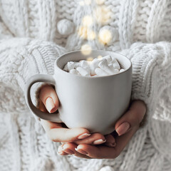 Fototapeta na wymiar Female hands hold a mug of coffee, cocoa with marshmallows. Woman in white knitted sweater, fall winter concept. Life style. Place for your text.