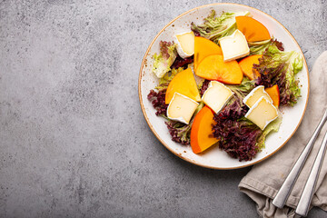 Top view flat lay persimmon salad with brie cheese, fresh salad leaves on white plate stone gray background, seasonal fruit salad as appetizer, vegetarian healthy food space for text