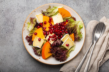 Top view flat lay persimmon salad with brie cheese, fresh salad leaves, pomegranate seeds on white plate and stone gray background, seasonal fruit salad as appetizer, vegetarian healthy food