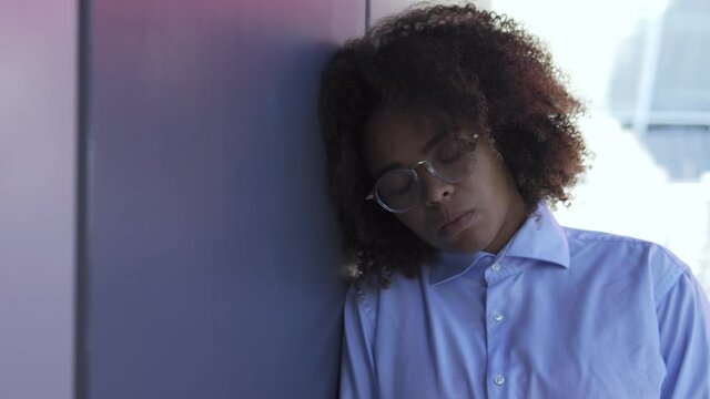Deeply upset frustrated black woman with afro-hair in stylish eyeglasses leaning on wall hiding in office hall, suffering bullying or racial discrimination, sexual harassment at work, abuse