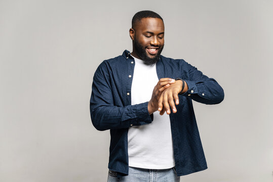 Positive African-American Guy Checking Notification On The Screen Of Smart Watch, Standing Isolated On Gray, Smiling Black Man In Casual Wear Looks At Smart Band On His Wrist, Controls Time Or Rate