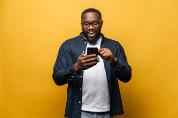 Portrait of cheerful young black guy sending message on mobile phone against yellow background. Happy African-American man using smartphone, enjoying chatting online, messaging in social networks