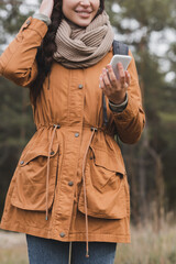 cropped view of smiling woman in autumn clothes holding mobile phone during walk in forest