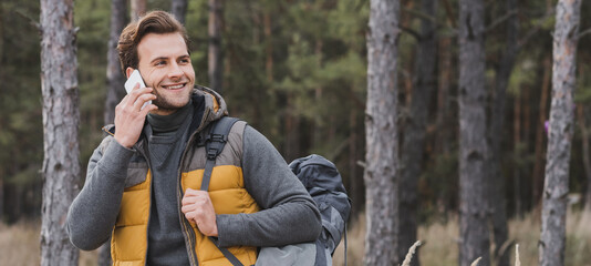 smiling tourist talking on mobile phone while walking in autumn forest, banner