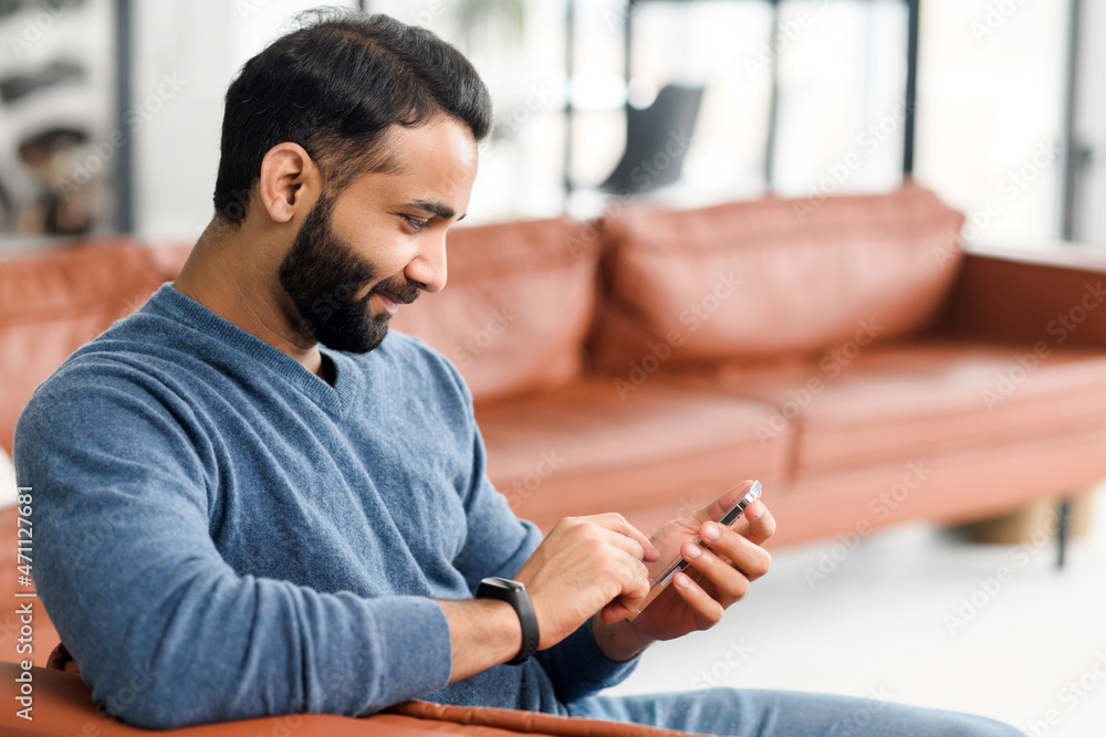 Fototapete handsome indian man sending messages on the smartphone, sitting on the comfortable couch with a cell