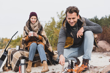 pleased woman sitting with thermo cup under warm blanket near boyfriend and bonfire