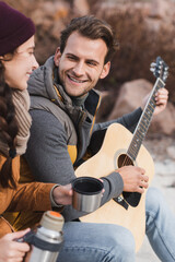 happy man playing acoustic guitar to blurred woman with thermos