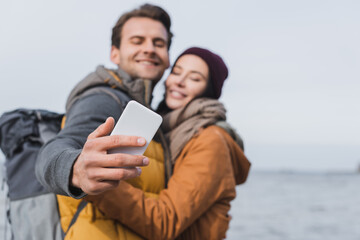 blurred couple in autumn clothes taking selfie on mobile phone while walking near river