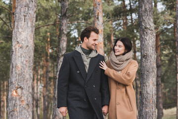 cheerful woman touching shoulder of smiling boyfriend while walking in autumn park