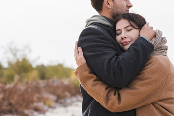 pleased woman with closed eyes hugging man in autumn coat outdoors