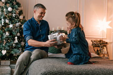 A young father gives Christmas gifts to his smiling daughters near a decorated Christmas tree. children's joy and delight