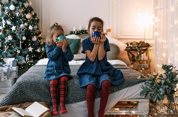 two little girls drinking tea on the bed against the background of a Christmas tree