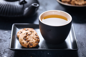 Dark grey cup of tea with cookies and teapot in background in a dark moody atmosphere 