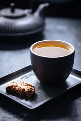 Dark grey cup of tea with cookies and teapot in background in a dark moody atmosphere 