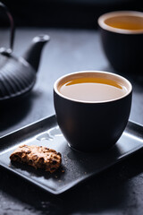 Dark grey cup of tea with cookies and teapot in background in a dark moody atmosphere 