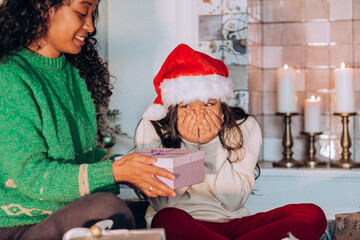 Curly African American woman and brunette long-haired daughter wearing holiday hats open present box sitting against Christmas tree