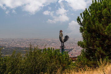 Denizli, Turkey - August 27 2021: The giant metal rooster, which was built in the urban forest and is the symbol of Denizli City with panoramic city view.