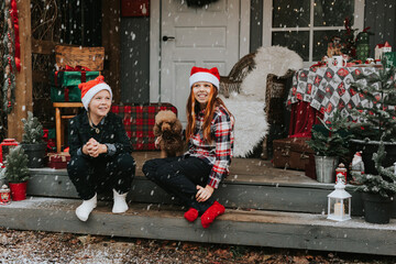 siblings teenage girl sister and cute boy brother in knitted sweater and hat having fun with first snow at porch of country house, concept of winter sports and Christmas holidays for children outdoor