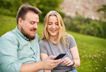 Young millennial couple checking social media and laughing together on a funny video in a park - Teenagers sharing a story on web - Modern technology and people lifestyle concept