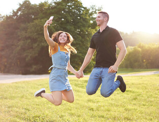 Millennial couple holding each other's hand and jumping together outdoors in a park while taking selfie - Happy people lifestyle concept