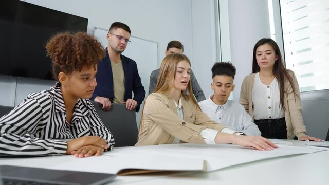 Young architects discussing masterplan lying on table, woman in center showing something on paper with hands, her multiethnic colleagues sitting beside and standing behind. Concept of business
