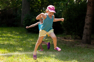 Two active joyful children, girls playing with water in the backyard jumping, running around, sisters siblings summer leisure, water hose, having fun outdoors, leisure, recreation, togetherness