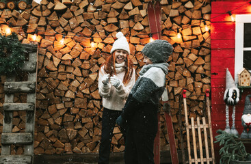 Fototapeta na wymiar siblings teenage girl sister and cute boy brother in knitted sweater and hat stand at porch of country house with sleds and skates, concept of winter sports and Christmas holidays for children outdoor