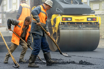 A team of workers with shovels in special yellow clothes is working on the road and laying new asphalt. Repair of asphalt concrete pavement. Workers stand next to a roller asphalt stacker