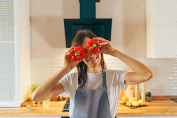 Young happy woman hold red peppers near eyes like glasses in modern cozy kitchen interior, fun...