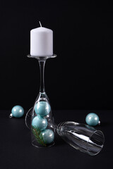 White candle on the stem of a champagne glass with blue christmas balls and a glass beside on a black background. The concept of a candlestick in the interior for the new year. Vertical photo